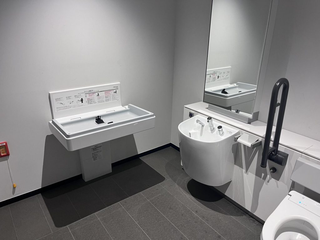 車椅子使用者対応トイレの壁面に取り付けられた、折りたたみ式のおむつ交換台(ベビーベッド)。手前に開いた状態で、右奥にオストメイト用設備と手すりがある。