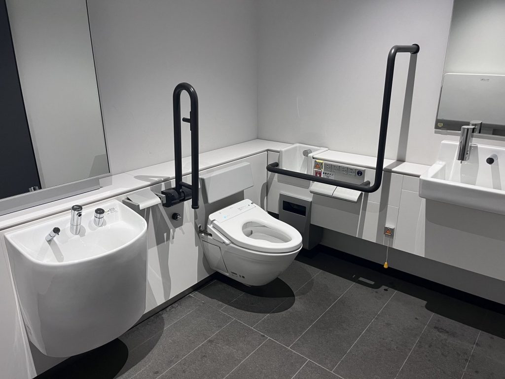車椅子使用者対応トイレの内部。手すりを備えた便器の左手前にオストメイト用の水栓器具付き汚物流しがあり、奥に洗面台がある。