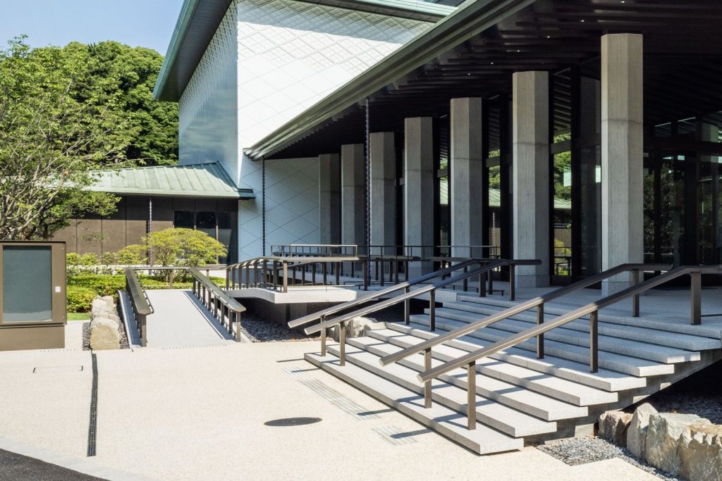 建物入口の階段と、その左側に設けられた手すり付きのスロープ。スロープは折り返しながら入口へ向かって上がっている。